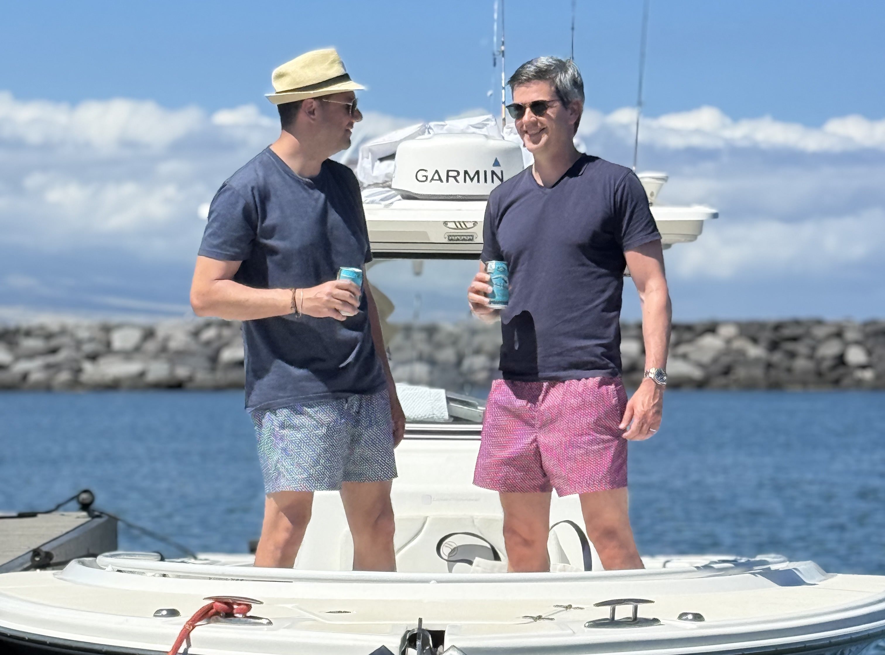 Two men standing on a white boat deck outdoors by the water, holding drinks. One man wears a beige hat, a navy T-shirt, and light patterned shorts; the other wears a dark navy T-shirt and pink shorts.
